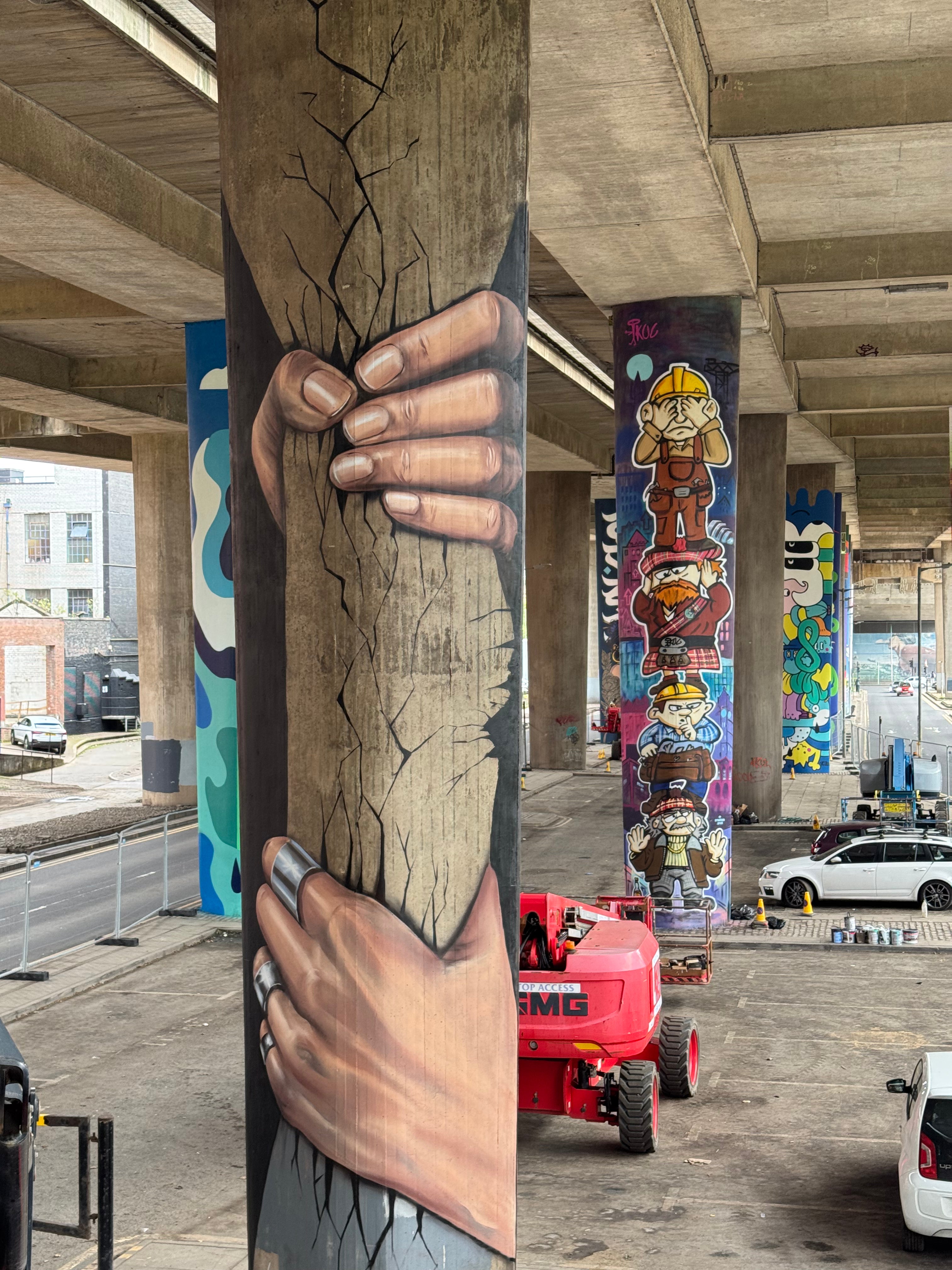 A large mural painted on kingston bridge pillar shows two hands gripping a cracked tree trunk. Other colorful graffiti murals are visible on adjacent pillars, with a red paint cart and parked cars in the background.